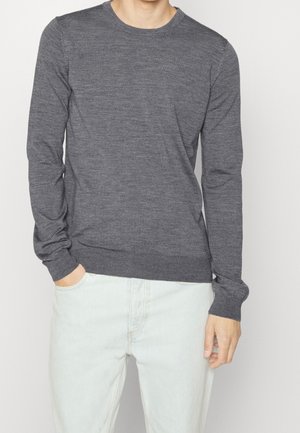 Mann trägt einen eng anliegenden dunkelgrauen Langarmpullover und hellblaue Jeans, eine Hand in der Hosentasche, die andere locker an der Seite.