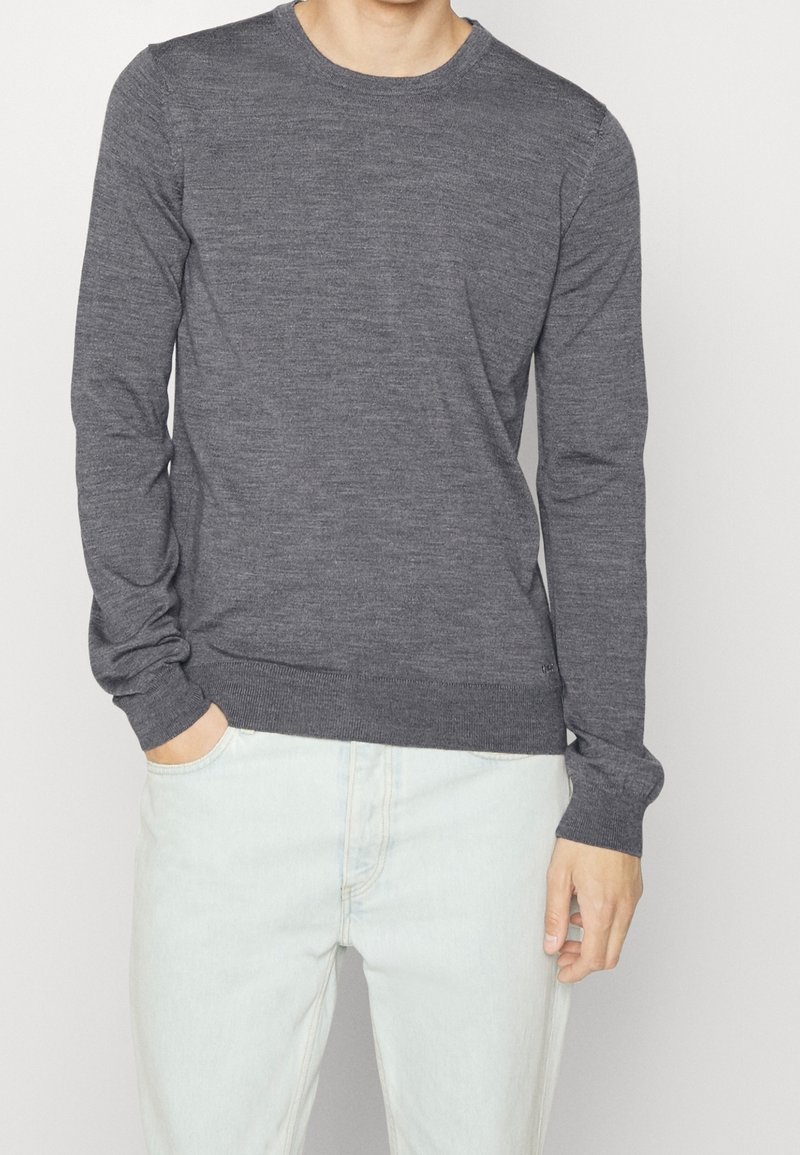 Homme portant un pull ajusté à manches longues gris foncé et un jean bleu clair, une main dans la poche et l’autre détendue sur le côté.