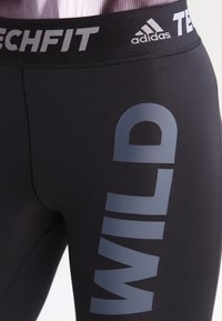 Mustat legginssit, joissa on sileä ja joustava pinta, suurella harmaalla "WILD" -grafiikalla sivussa ja joustovyötäröllä, jossa on Adidas-brändäys.
