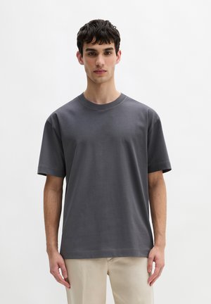 Jonge man met kort donker haar, gekleed in een effen donkergrijs T-shirt met korte mouwen en lichtbeige broek, staand tegen een witte achtergrond.