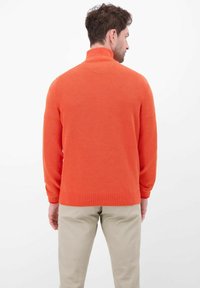 Oranger gestrickter Pullover mit hohem Kragen, strukturiertem Muster und langen Ärmeln; kombiniert mit hellfarbenen Hosen.