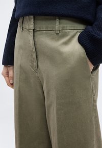 Κοντινό πλάνο ελιάς πράσινου παντελονιού με πλαϊνές τσέπες, θηλιές για ζώνη, και ένα άτομο που φοράει σκούρο μπλε πουλόβερ με το ένα χέρι στην τσέπη.