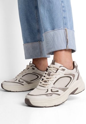 Pieds portant des baskets en mesh beige et gris avec des semelles épaisses et un jean bleu clair remonté sur fond blanc.