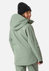 Jasnozielona wodoodporna kurtka z kapturem, zapinanymi kieszeniami i czarnymi akcentami na mankietach; noszona na dopasowanym stroju.