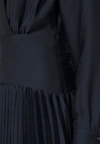 Robe en tissu texturé foncé avec une large ceinture ajustée, des manches longues et un détail de jupe plissée à la taille.