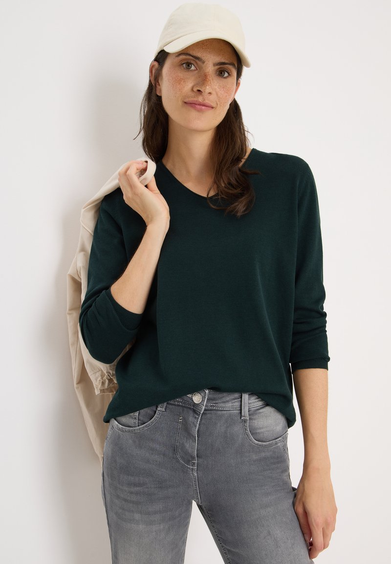 Dunkelgrüner Pullover mit Rundhalsausschnitt, enganliegende Ärmel, getragen über hellgrauen Jeans. Eine hellfarbige Mütze rundet das Outfit ab.