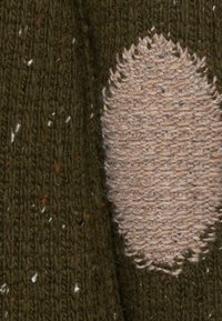 Patch en tricot présentant un tissu texturé de couleur beige clair avec une forme ovale irrégulière, sur un fond vert foncé parsemé de touches colorées.