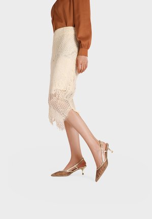 Donna che indossa una gonna midi in pizzo crema, una camicetta marrone a maniche lunghe e décolleté slingback con tacco basso e motivo dorato, in piedi su sfondo bianco.