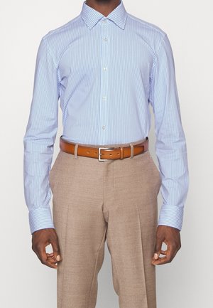 Homme portant une chemise à rayures bleu clair et blanc rentrée dans un pantalon beige avec une ceinture en cuir marron.