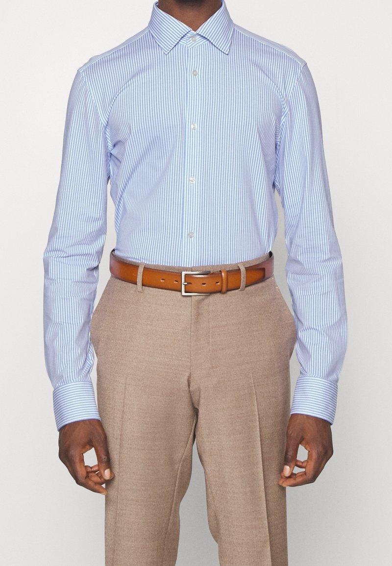 Homme portant une chemise à rayures bleu clair et blanc rentrée dans un pantalon beige avec une ceinture en cuir marron.