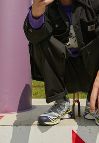 Persona accovacciata su un blocco di cemento, indossa pantaloni neri, un maglione viola, una giacca nera e scarpe da corsa grigie con dettagli gialli e blu.