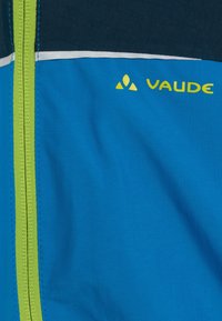 Vaude-jacka med lysande blått tyg, kontrasterande marinblått, reflexdetaljer, grön dragkedja och logotyp i gult. Strukturerad yta.