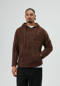 Hoodie en polaire marron à coupe décontractée, avec capuche à cordon, poche kangourou et surface texturée. Porté sur une chemise blanche et un pantalon noir.