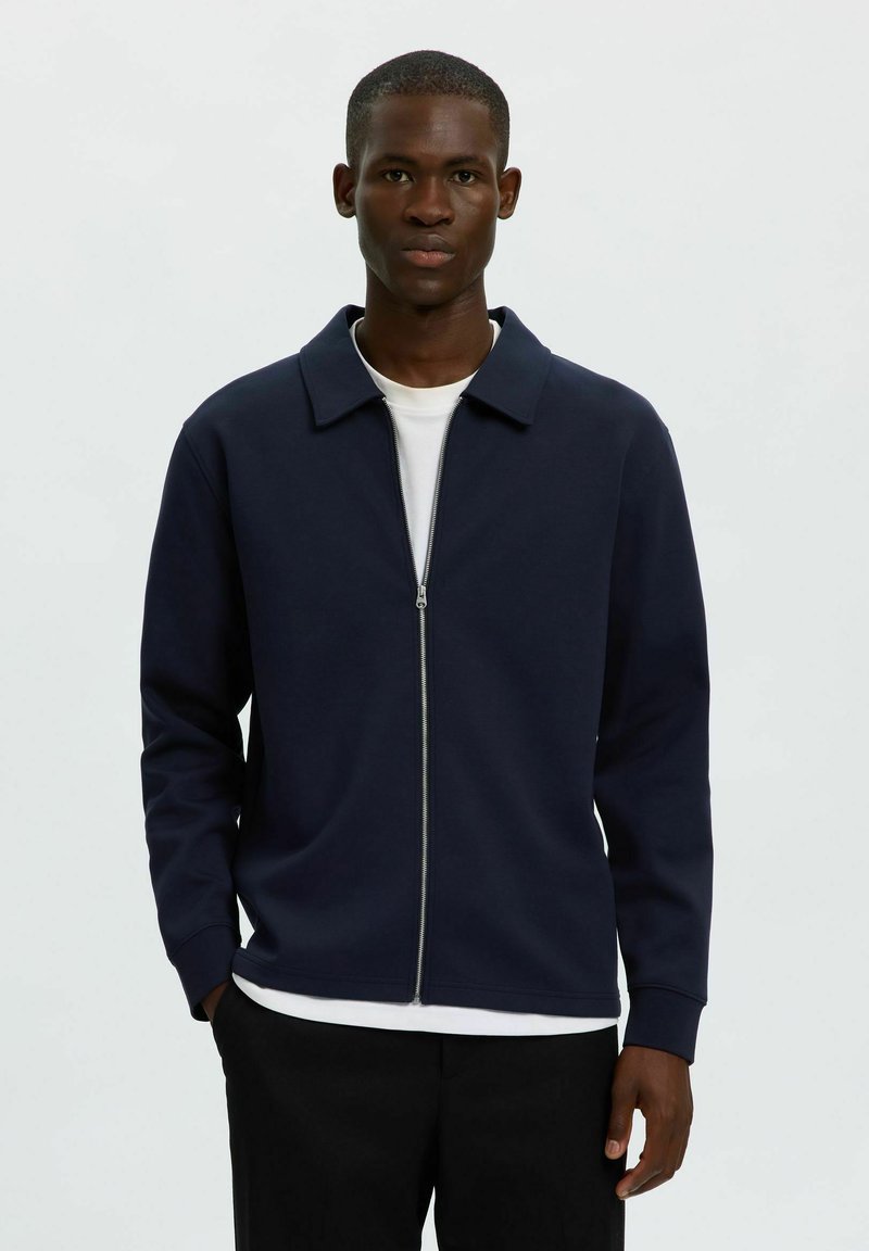 Jeune homme portant une veste zippée bleu marine sur une chemise blanche et un pantalon noir, debout les mains dans les poches devant un fond blanc uni.