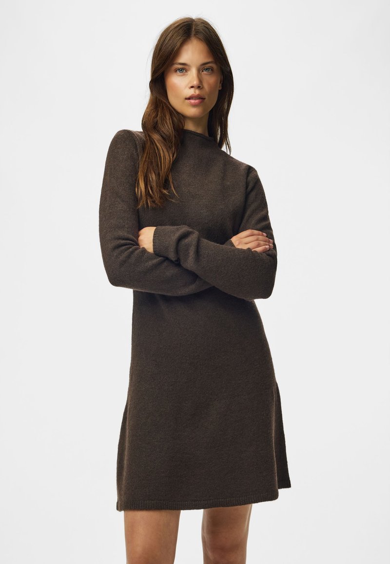 Femme aux longs cheveux bruns portant une robe en tricot marron foncé à manches longues, debout, les bras croisés, devant un fond uni.