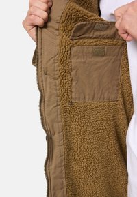 Veste zippée marron avec un intérieur moelleux, présentant un tissu extérieur lisse, une doublure douce et texturée, et une petite poche à l'intérieur.