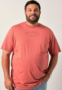 Korálové tričko vyrobené z měkké látky, s krátkými rukávy a kulatým výstřihem. Na přední straně je nápis "MONTREAL Northern Roots" v kontrastní barvě.