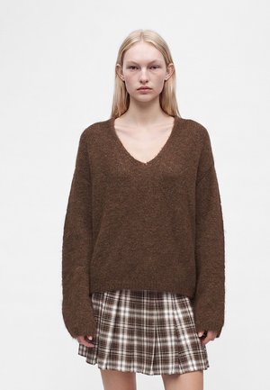 Ung kvinde med glat blondt hår iført en brun, blød V-hals sweater og et brun-hvid ternet plisseret nederdel.