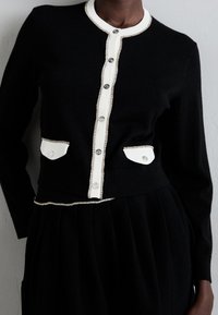 Svart kavaj med vit kant och silverknappar, med två små vita knappfickor, bärs med en plisserad svart kjol.