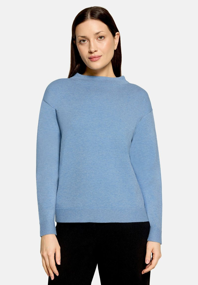 Femme aux cheveux raides et foncés portant un pull bleu clair uni à manches longues et un pantalon noir sur un fond clair.