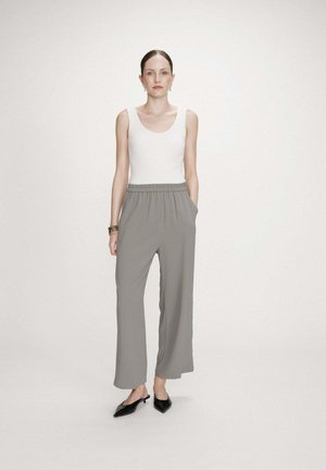 Femme debout les mains dans les poches, portant un haut blanc sans manches, un pantalon gris à jambes larges, des chaussures noires à bout pointu et des boucles d'oreilles.