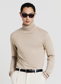 Beiger Rollkragenpullover mit gerippter Textur und figurbetontem Design, kombiniert mit einem schwarzen Gürtel und weißen Hosen; dunkle quadratische Sonnenbrille als Accessoire.