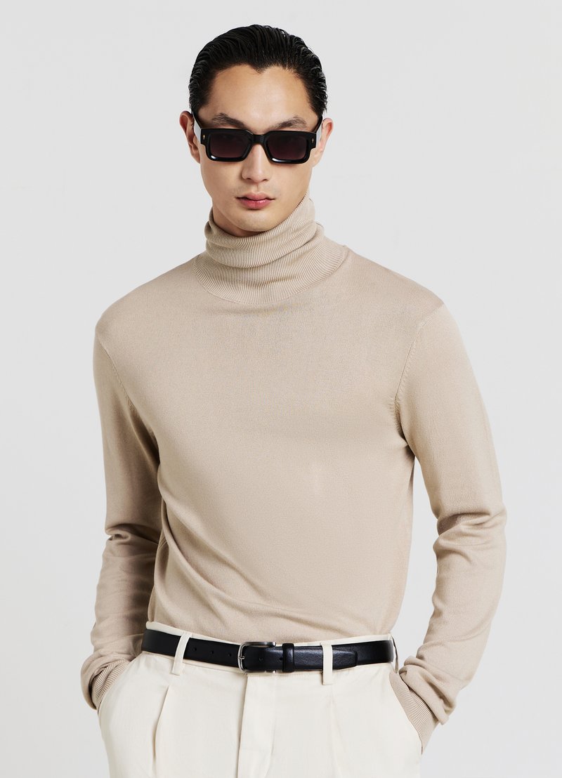 Beiger Rollkragenpullover mit gerippter Textur und figurbetontem Design, kombiniert mit einem schwarzen Gürtel und weißen Hosen; dunkle quadratische Sonnenbrille als Accessoire.