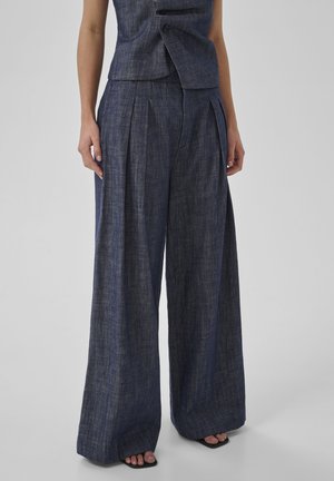 Kamizelka bez rękawów na guziki w zestawieniu z wysokim stanem, szerokimi spodniami z ciemnego denimu, noszona z czarnymi sandałami na otwartym palcu.