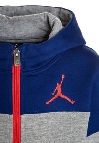 Sweat à capuche bleu et gris, avec une fermeture éclair rouge et un logo Jumpman brodé. Le matériau est doux, avec une doublure intérieure contrastante.