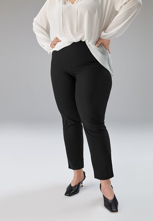 Schwarze, eng anliegende Hose mit glatter Stoffoberfläche, kombiniert mit einer weißen Bluse, die locker sitzt und einen Kordelzug am Ausschnitt hat, um die Taille zu betonen.