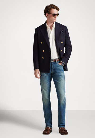 Jauns vīrietis ar saulesbrillēm, tumszilu uzvalka žaketi ar zelta pogām, baltu kreklu, zilajām džinsu biksēm, brūnu jostu, pulksteni un brūniem bārkstainiem mokasīniem stāv.