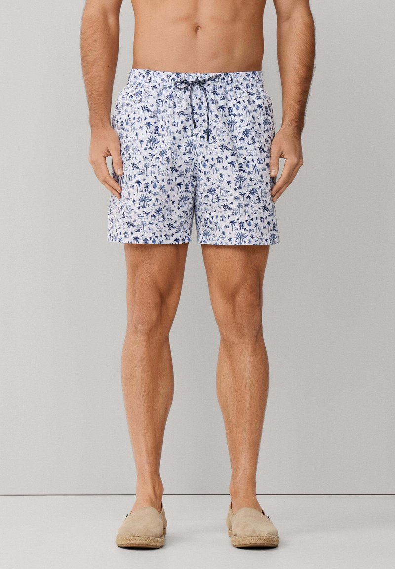 Männliches Modell trägt weiße Badeshorts mit blauem tropischem Muster und beige Slip-on-Schuhe, steht vor einem einfachen grauen Hintergrund.