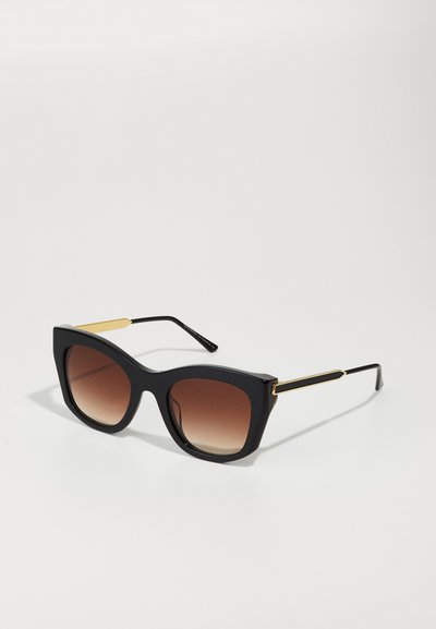 Black cat-eye sunglasses with a matte finish, gradient brown lenses, and gold-tone accents on the temples. Angular design and bold shape.