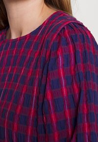 Roten und marineblauen karierter Bluse mit strukturiertem Stoff, die über plissierte Schultern und einen runden Ausschnitt verfügt. Auffälliges, gemustertes Design.