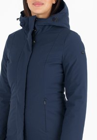 Veste isolante bleu marine avec capuche, dotée d'une fermeture éclair à l'avant, de boutons-pression et de deux poches latérales. Tissu lisse avec des détails de couture minimaux.