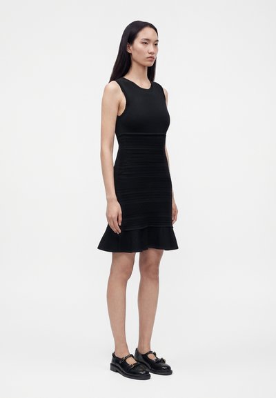 Femme aux longs cheveux noirs portant une robe noire sans manches avec une taille texturée et un ourlet à volants, debout les mains le long du corps, chaussée de chaussures noires.