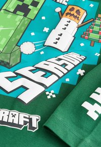 Zöld póló Minecraft mintával, pixelált karakterekkel, hópelyhekkel és szöveggel egy kék háttéren. Simogató, sima textúra.