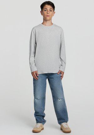 Camicia a maniche lunghe in cotone grigio con colletto tondo, abbinata a jeans in denim chiaro strappati e sneaker beige. Stile e vestibilità casual.