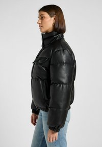 Chaqueta de plumón negra con un cuello alto, cuenta con un bolsillo frontal, puños acanalados y una textura brillante. El modelo lleva puesto unos jeans de color azul claro.
