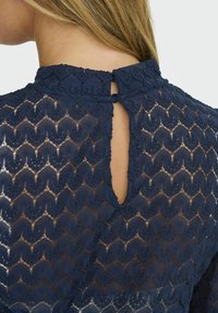 Top en dentelle bleu marine mettant en valeur un motif floral avec des bords festonnés et une ouverture en trou de serrure à l'arrière maintenue par un lien en boucle.