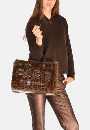Donna con un maglione marrone scuro e pantaloni metallici color bronzo, che tiene in mano una borsa di pelliccia con stampa leopardata su cui è ricamato in lettere grandi "PARIGI".