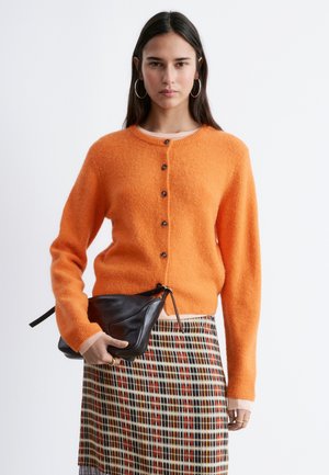 Vrouw draagt een oranje gebreid vest met knopen, ruitjesrok, hoepeloorbellen, en houdt een zwarte leren clutch vast tegen een effen witte achtergrond.