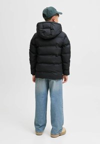 Doudoune noire avec capuche, présentant une texture matelassée et une coupe ample, associée à un jean large bleu clair et des bottes beiges.