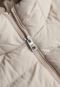 Veste matelassée beige avec une finition texturée, dotée d'une fermeture éclair métallique avec un tirette de marque et des détails matelassés.