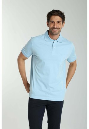 Homme souriant, portant un polo bleu clair à manches courtes et un pantalon sombre, debout avec les mains dans le dos sur un fond neutre.