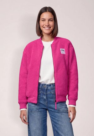 Jeune femme souriante portant une veste polaire rose vif, une chemise blanche et un jean bleu sur un fond clair uni.