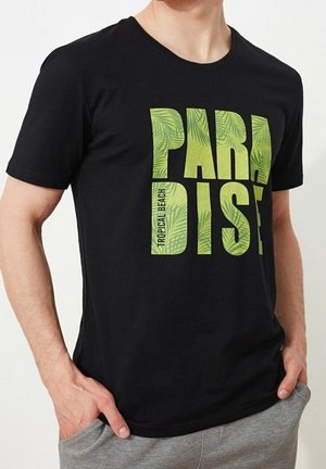 Mann trägt schwarzes T-Shirt mit grünem tropischem Blattdruck und graue Hose, Hände in den Taschen.