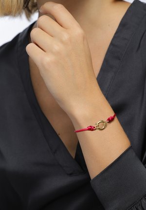 Poignet de femme portant un bracelet en cordon rouge avec un fermoir circulaire doré, main levée près du menton, blouse en satin noir visible.