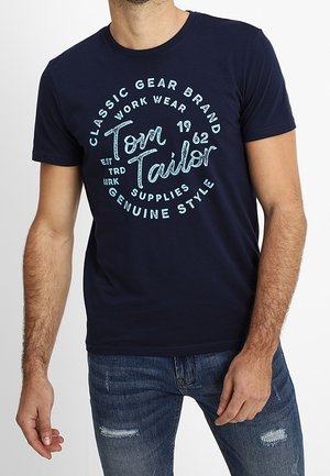 Marineblå bomulds-T-shirt med en cirkulær grafik i lyseblå med teksten "Tom Tailor, Classic Gear Brand, 1962." Rund halsudskæring.