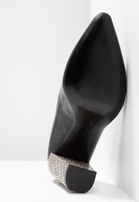 Escarpin noir en cuir avec un bout pointu et un talon en métal argenté texturé présentant un motif géométrique. Fini de surface lisse.